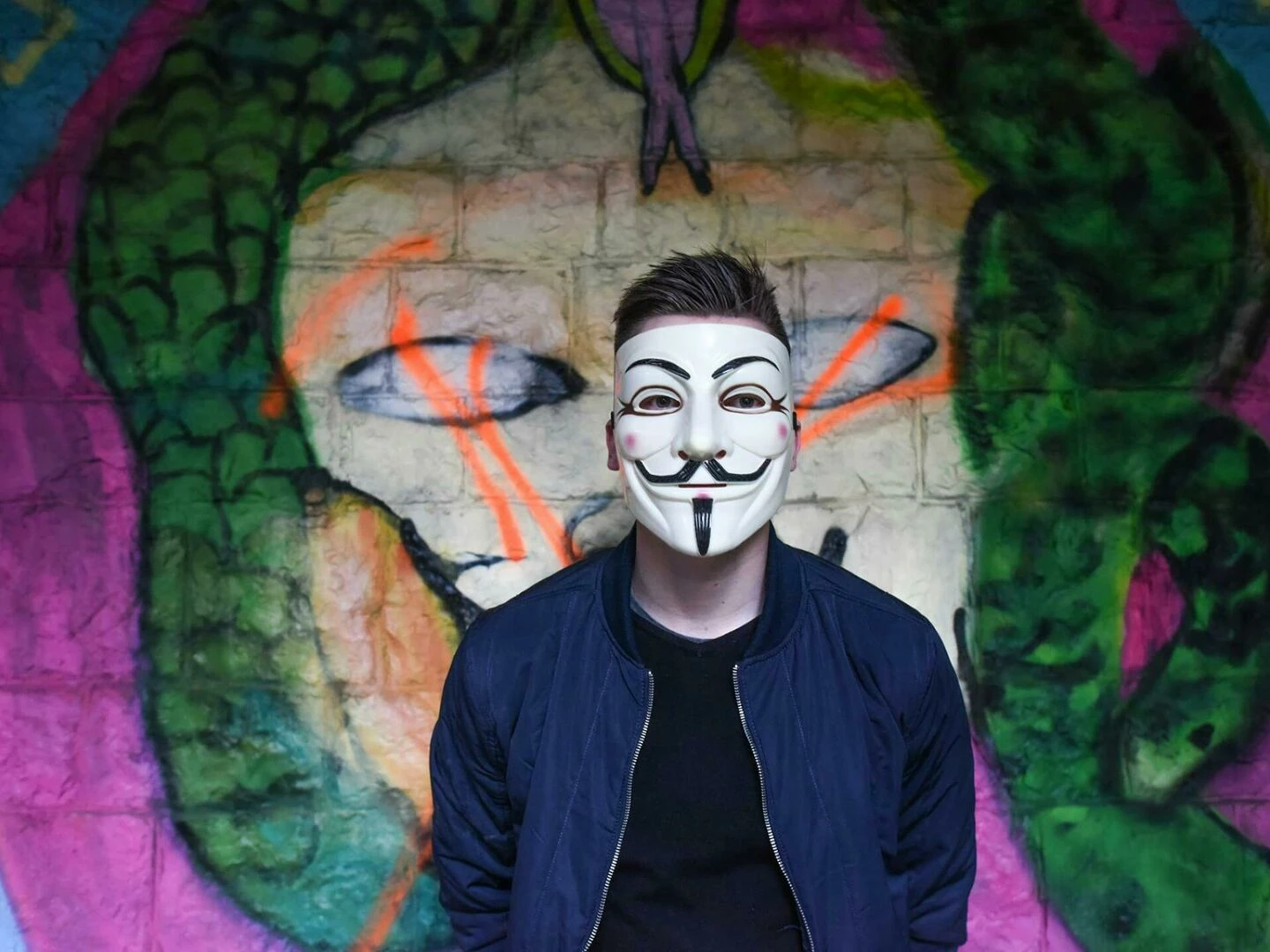 a person in a guy fawkes mask stands before colorful graffiti art creating an air of mystery