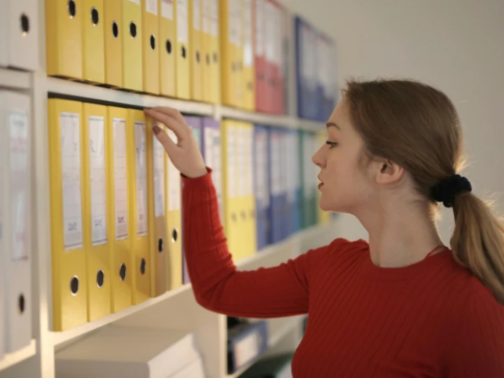 Side view of contemplating female assistant in casual style standing near shelves and choosing file with documents