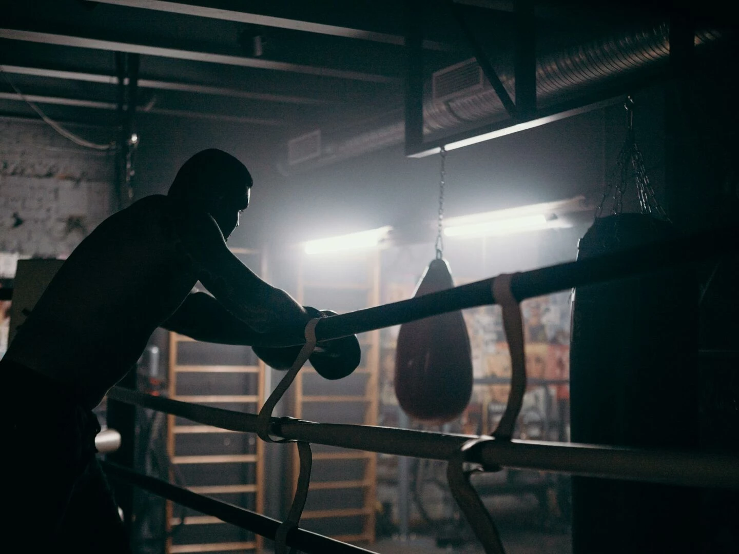 moody silhouette of a boxer leaning in a dimly lit gym with punching bag and boxing ring